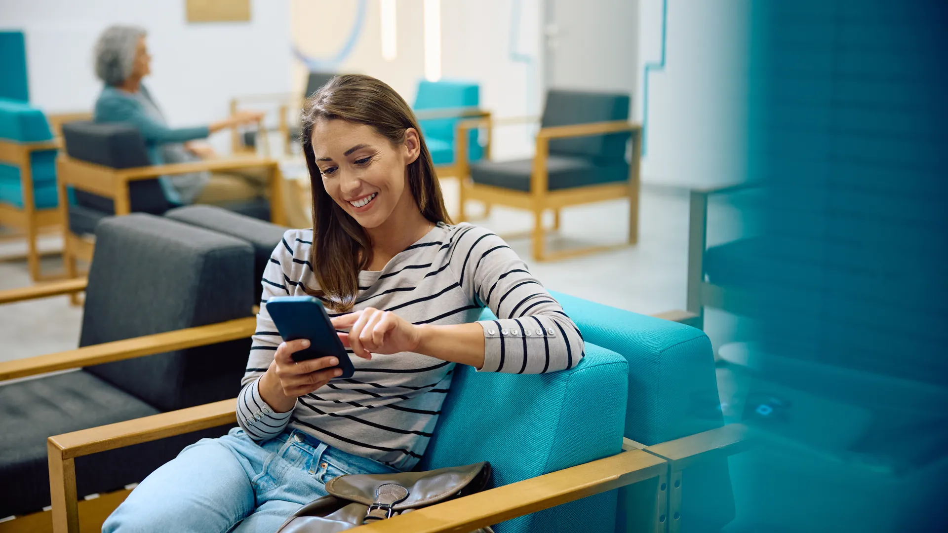 A smiling woman in a striped top looks at her phone while sitting on a turquoise chair in a modern waiting area.