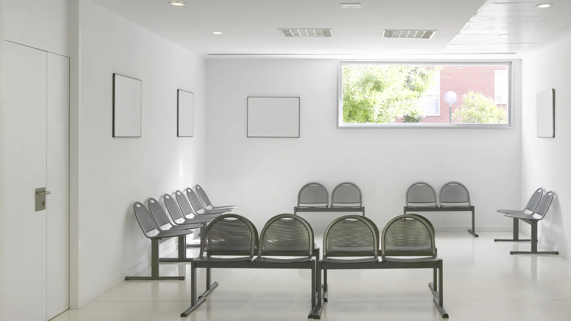 A bright, empty hospital waiting area with rows of grey metal chairs and a window.