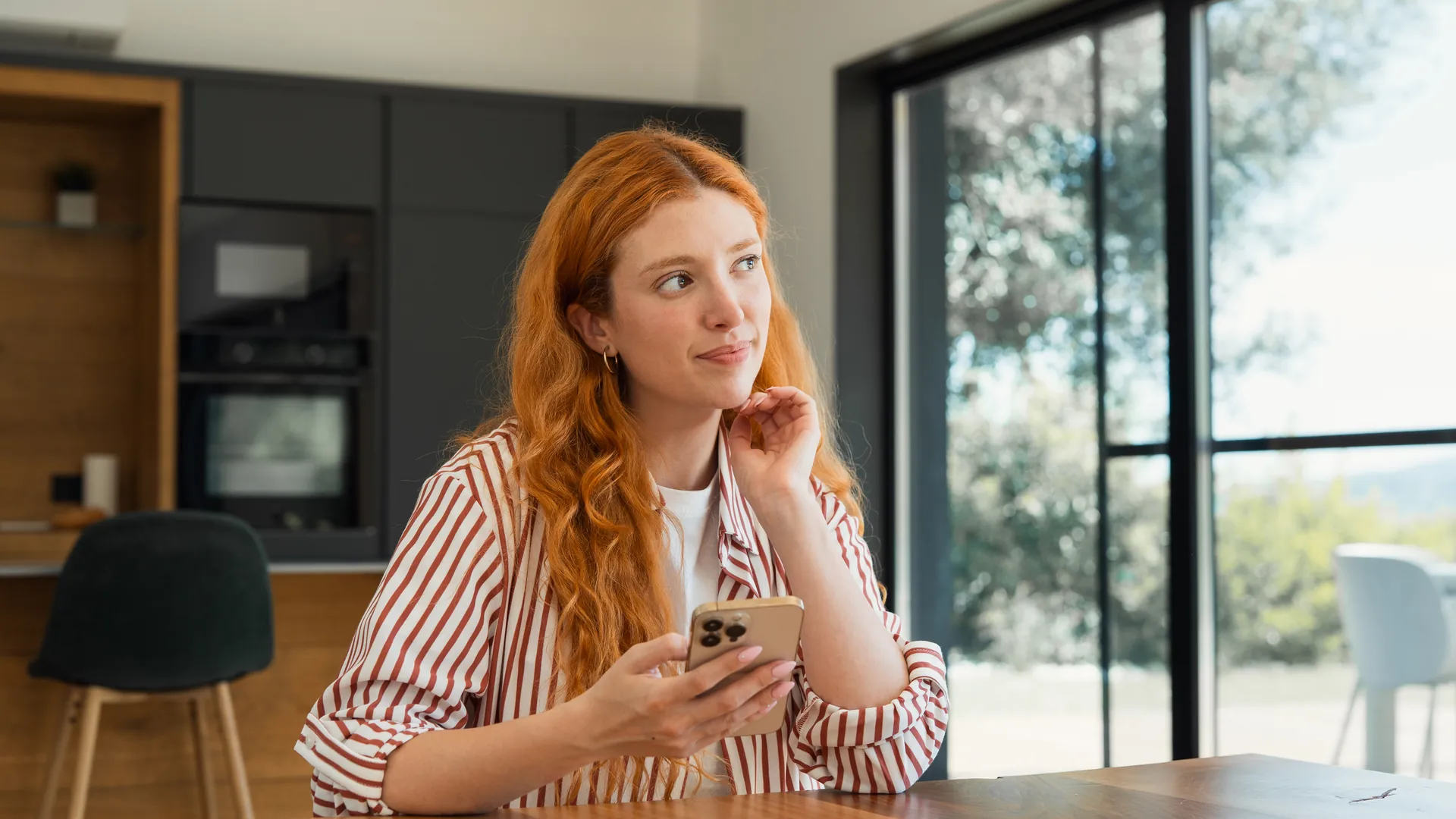 A red-haired woman in a striped shirt rests her chin on her hand, looking thoughtful, phone in hand, in a bright kitchen.