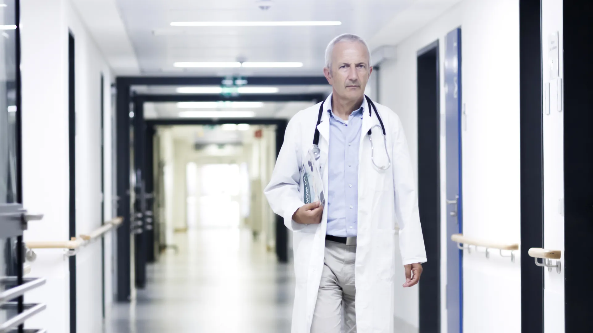 A grey-haired doctor in a white coat with a stethoscope walks down a long hospital corridor carrying papers.