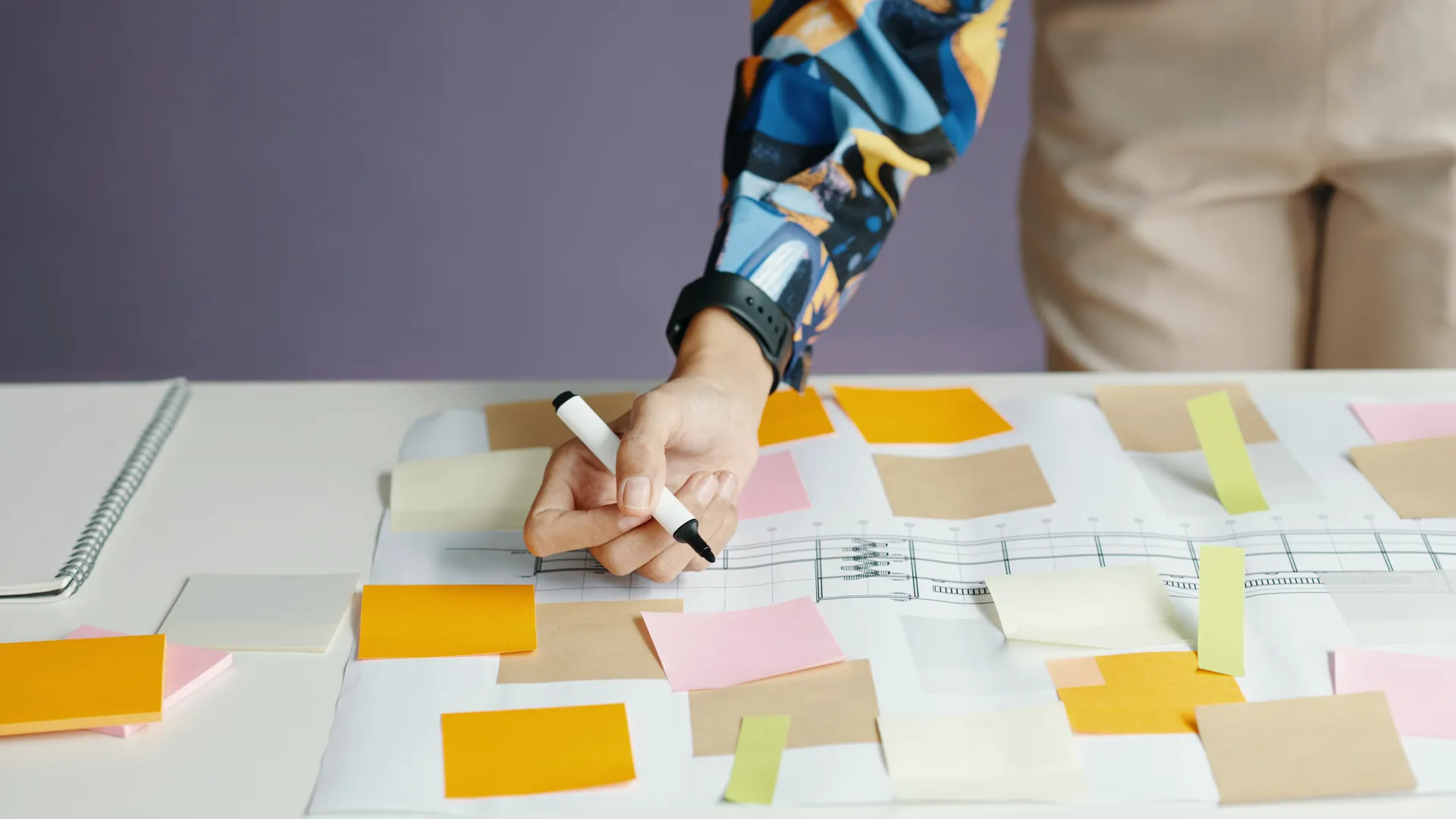 A person in a colourful patterned shirt writes on a table covered in sticky notes and a printed plan.