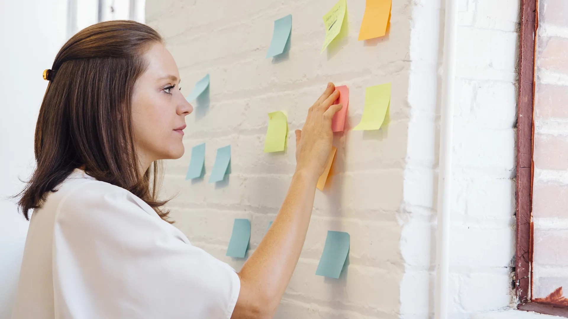 A woman in a white blouse presses coloured sticky notes onto a white brick wall while planning her ideas.