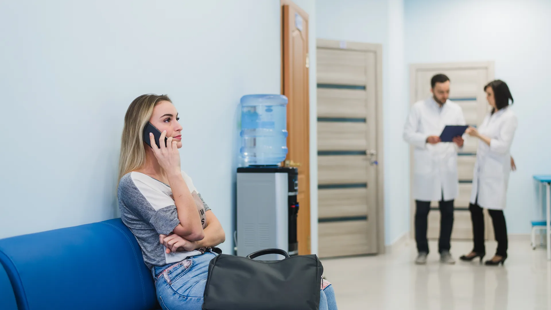 A woman sits on a blue bench in a waiting area, talking on her phone while two doctors chat behind her.