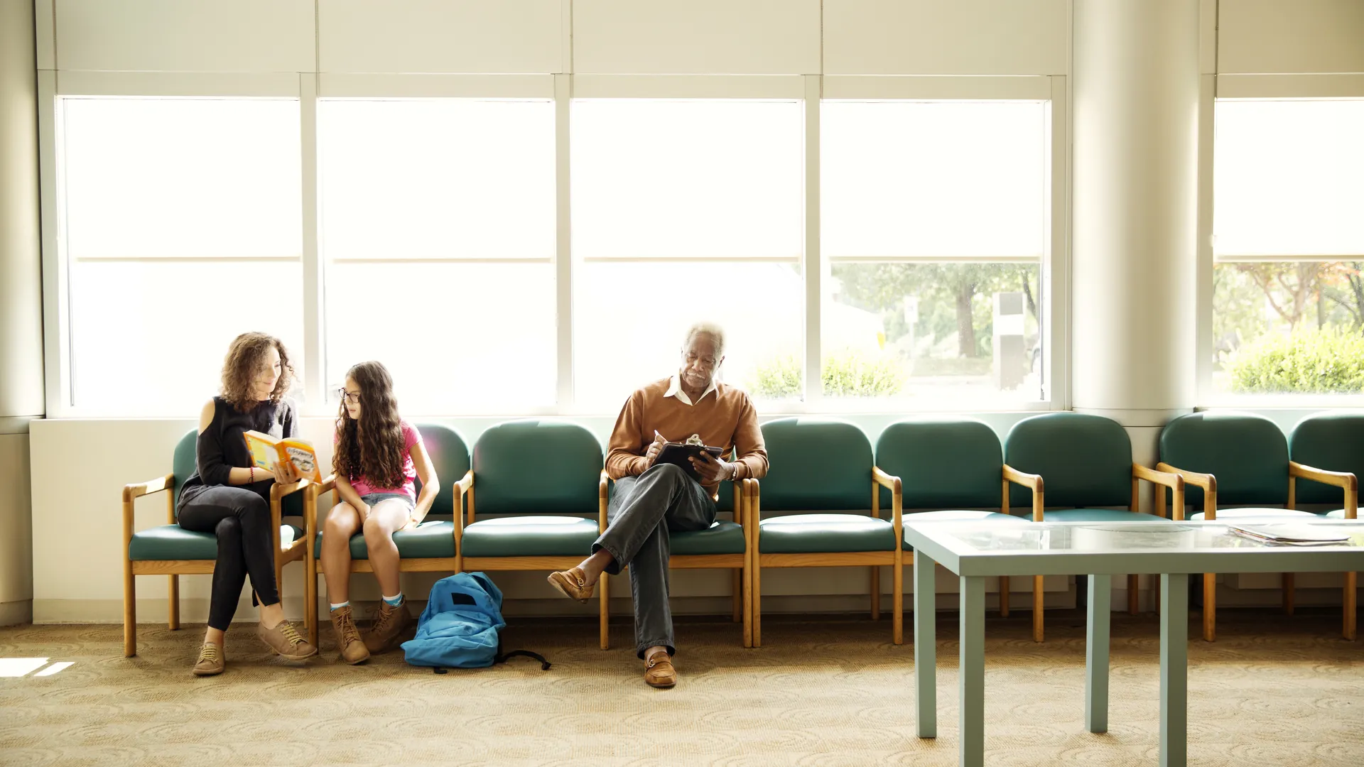 Patients sit on green waiting-room chairs along a wall of large windows in a bright clinic waiting area.