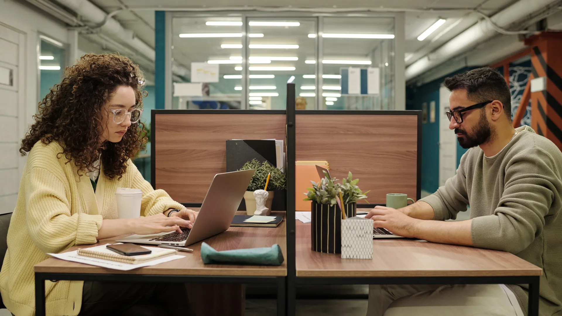 A woman in a yellow cardigan and a man in a green jumper work on laptops at desks in a modern open-plan office.