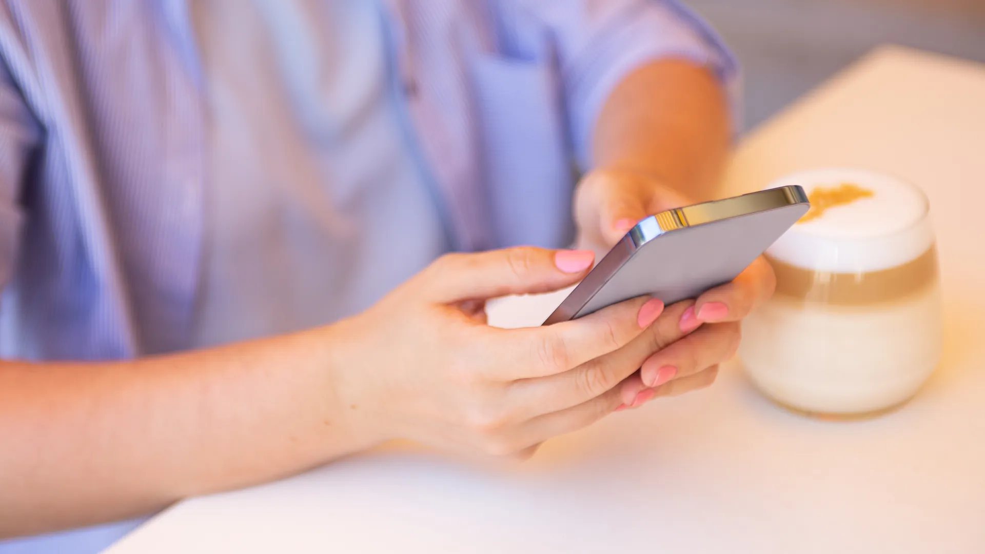 A woman in a lilac top holds a smartphone in both hands beside a cup of coffee.