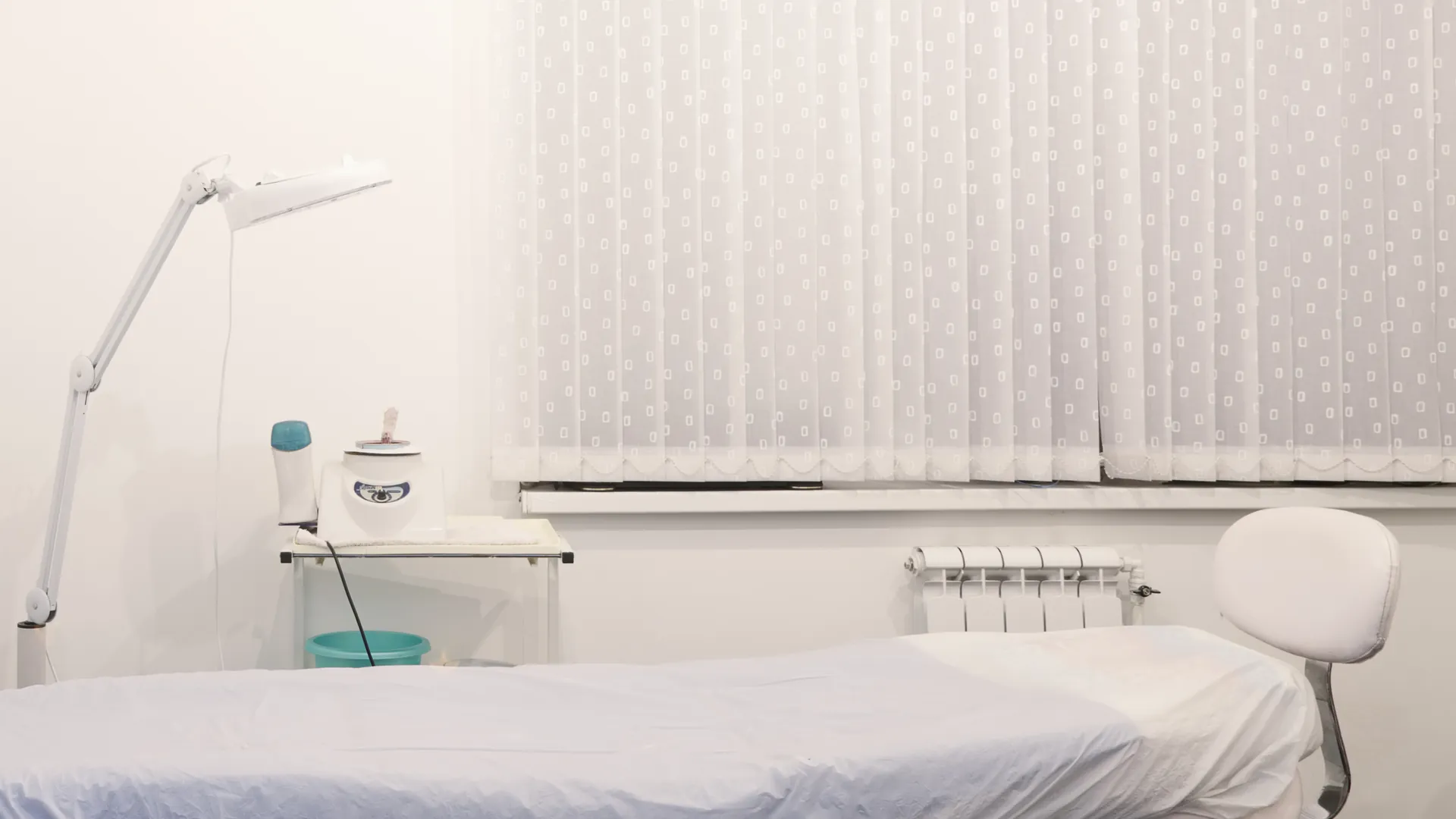 A clean treatment room with a white couch, an adjustable lamp and a wax heater beside a window with white blinds.