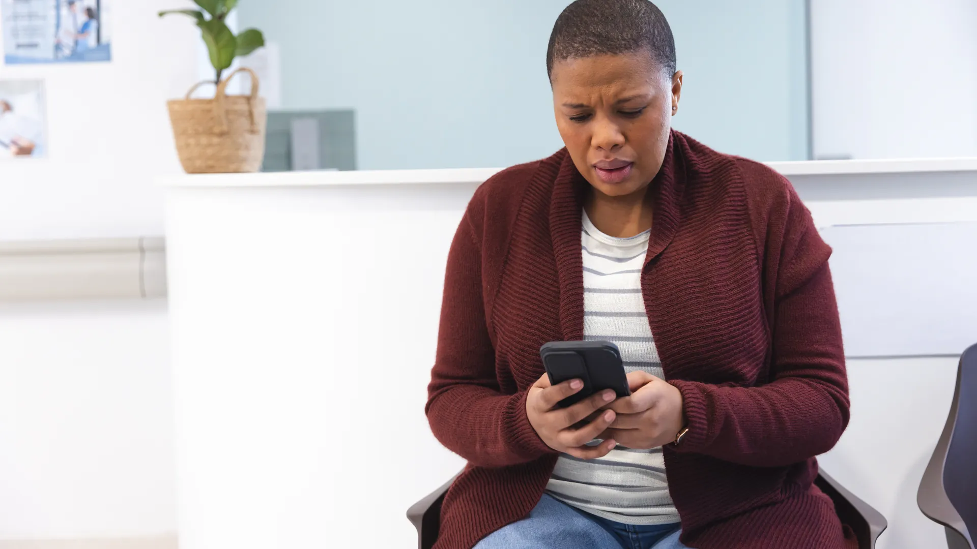A woman in a burgundy cardigan looks worried as she checks her phone in a bright waiting room.