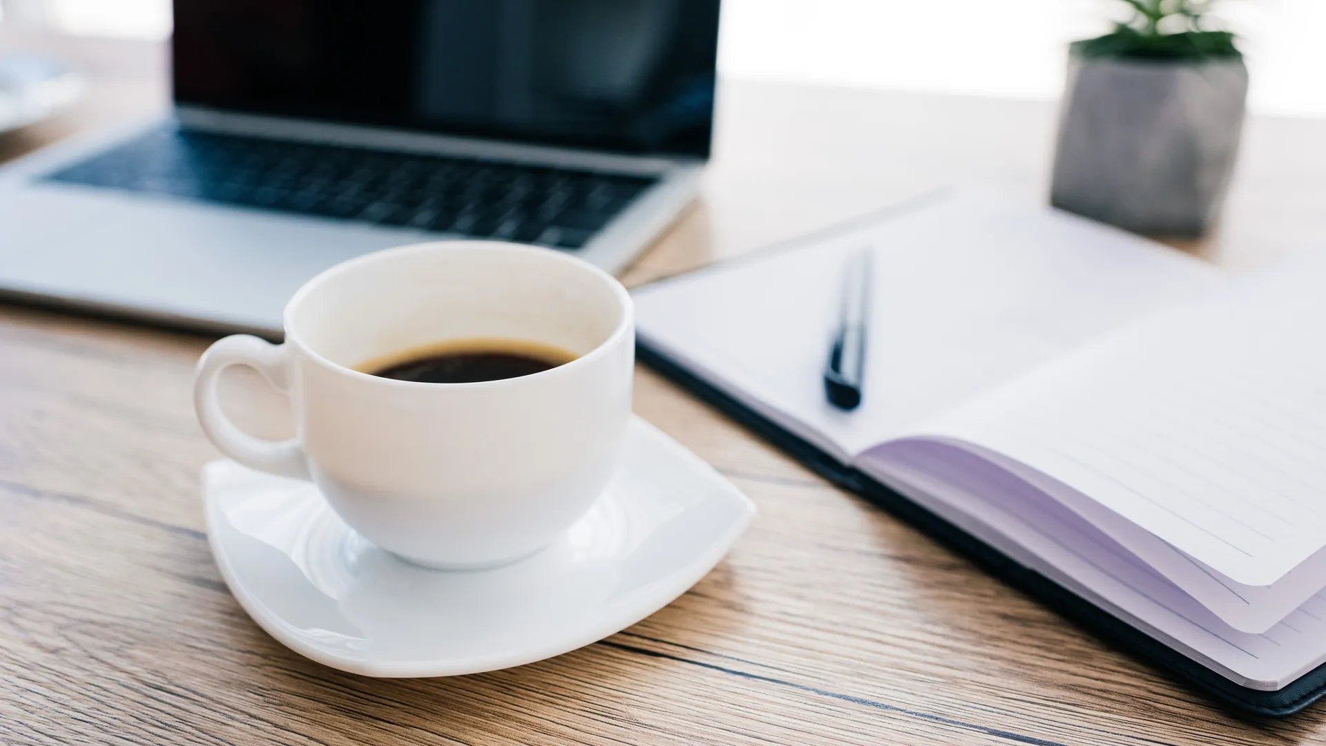 A cup of black coffee on a saucer beside an open notebook and a laptop on a light wooden desk.