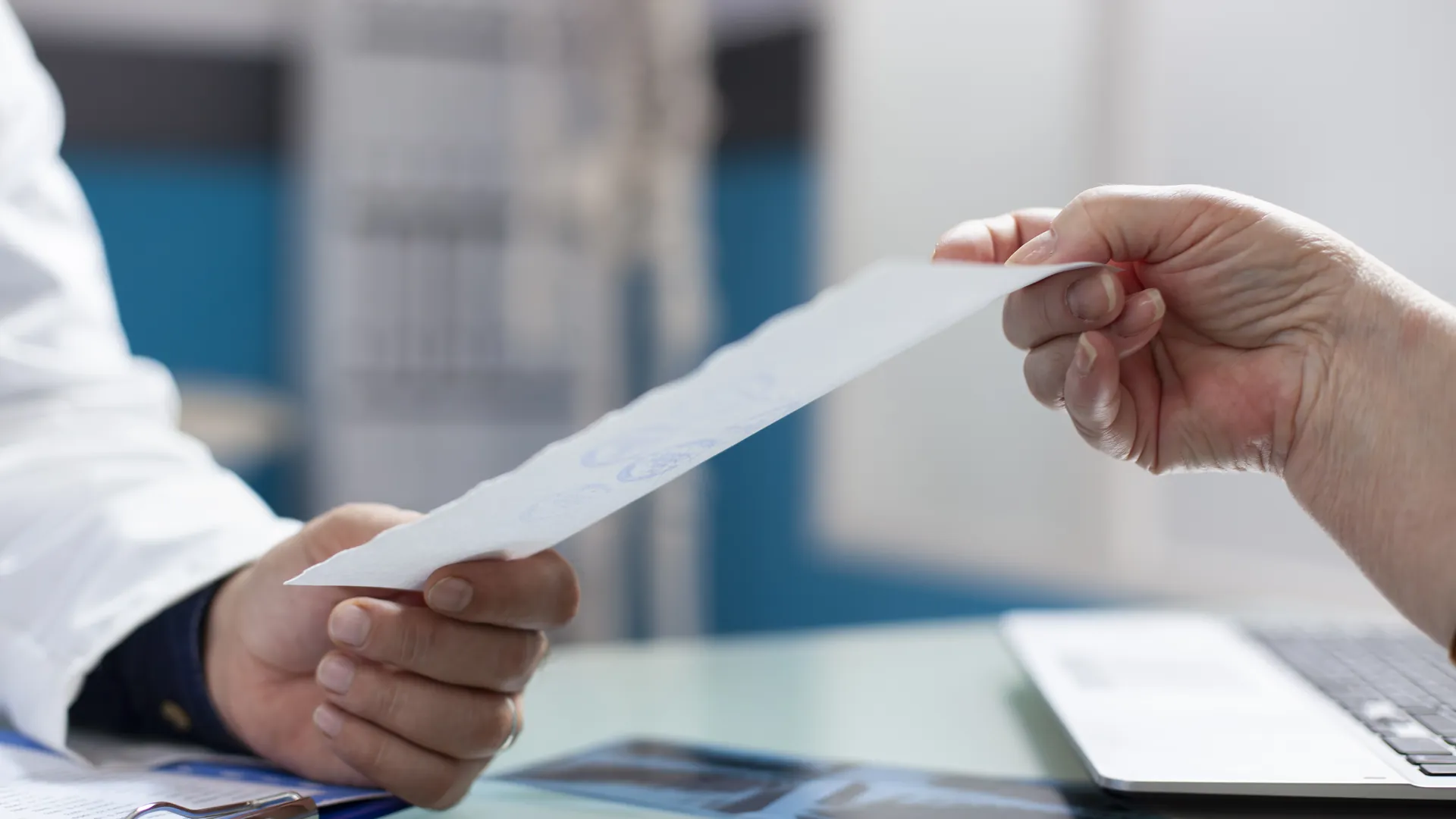 A doctor's hand passes a prescription slip across a desk to a patient's hand, with a laptop in the foreground.