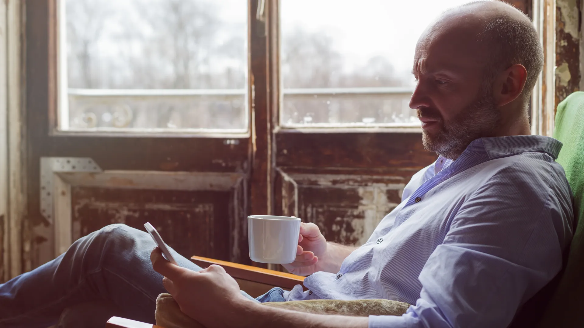 A man in a blue shirt relaxes with a cup of coffee and checks his phone beside a large old window.