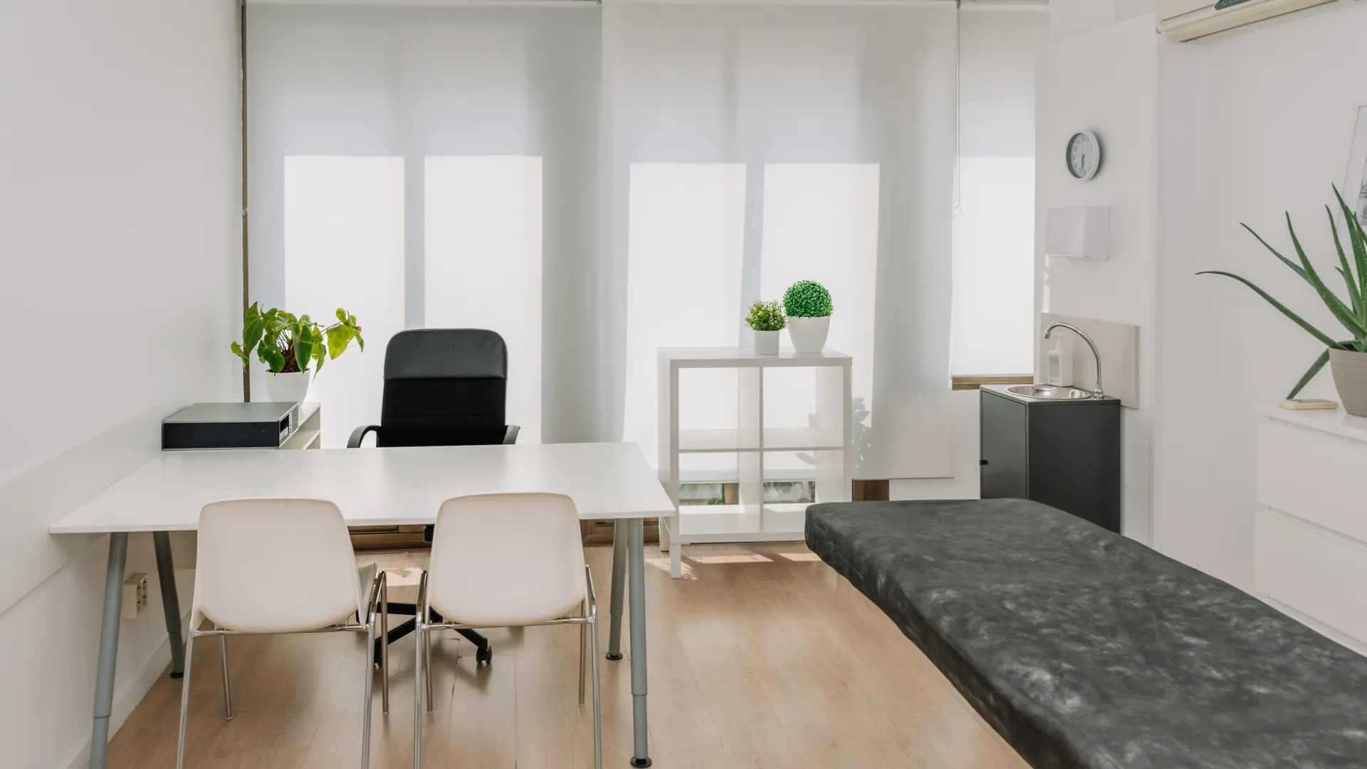 A bright consulting room with a white desk, two chairs, a black examination couch and a small plant by the window.