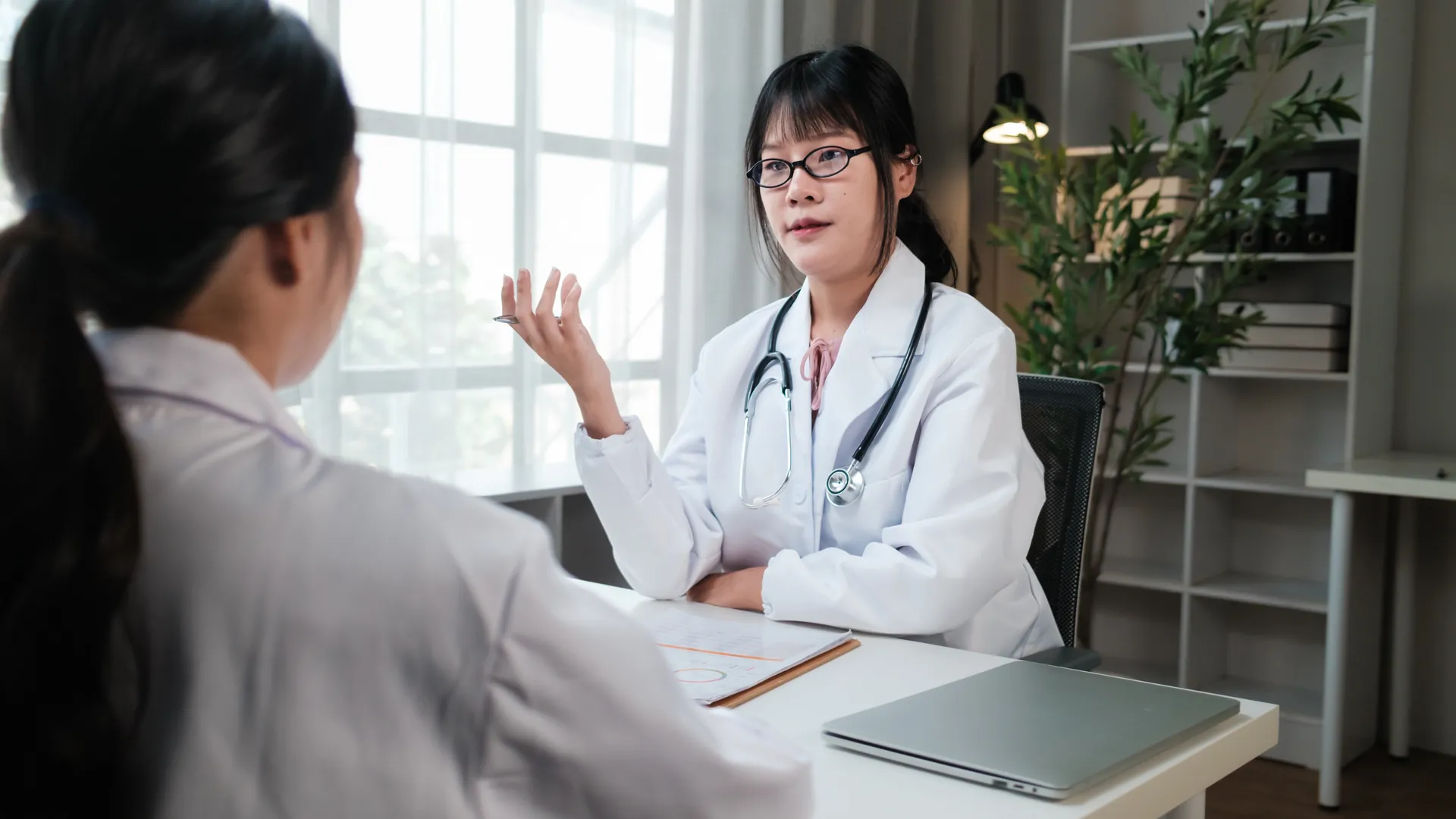 Two doctors in white coats sit at a desk in a bright office, one gesturing as she explains something to the other.