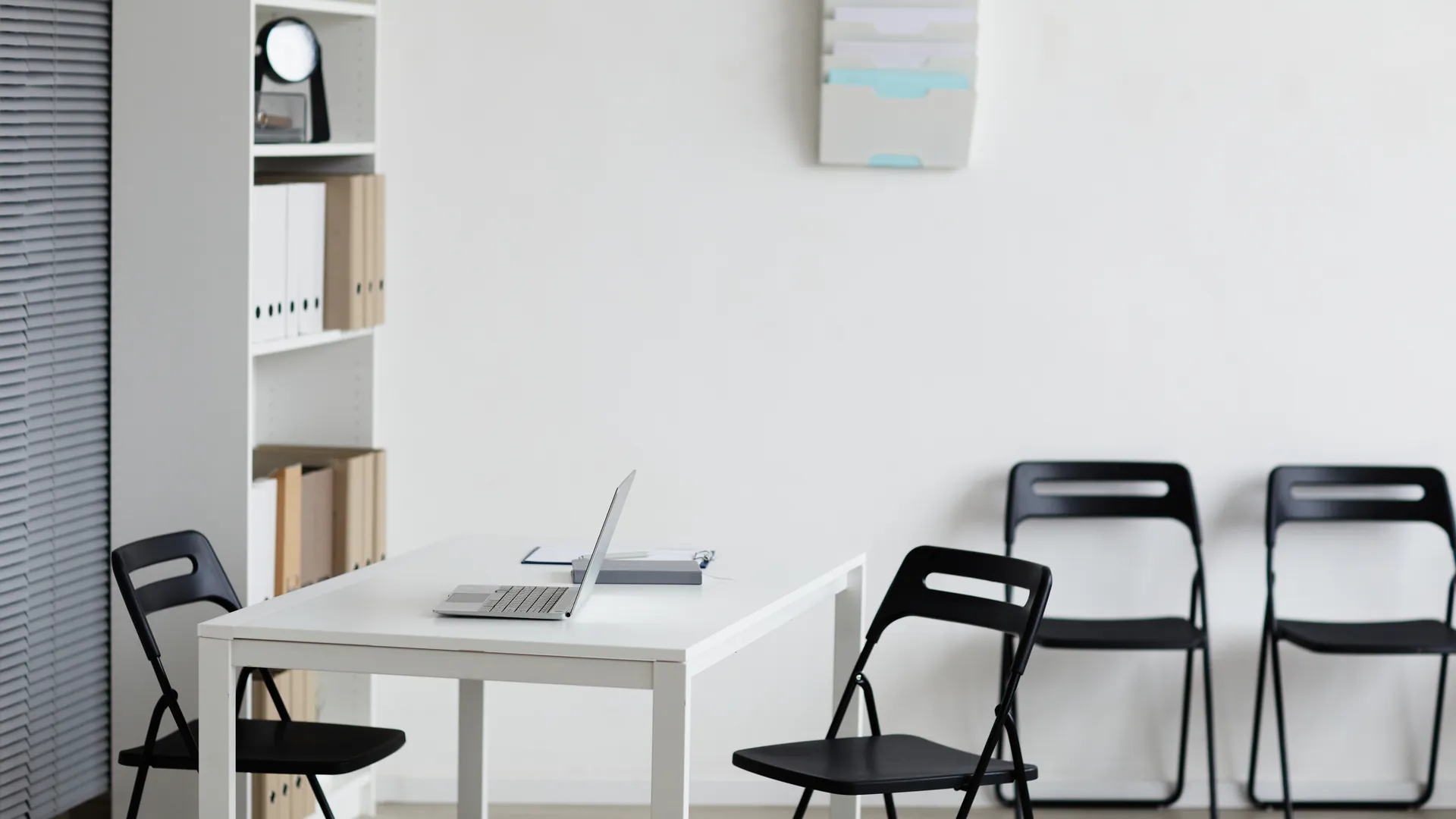 A white desk with a laptop, black folding chairs and shelves of folders in a bright, simple office room.