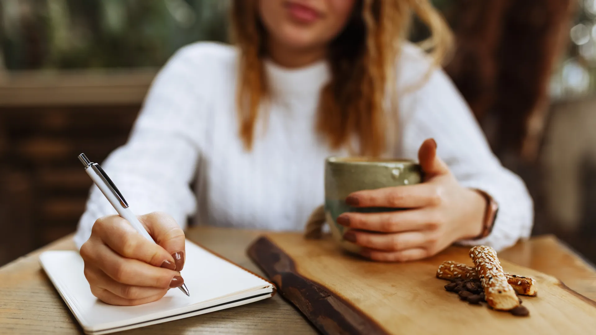 A woman in a white knit jumper writes in a notebook while holding a cup of coffee, with a snack on a wooden board.