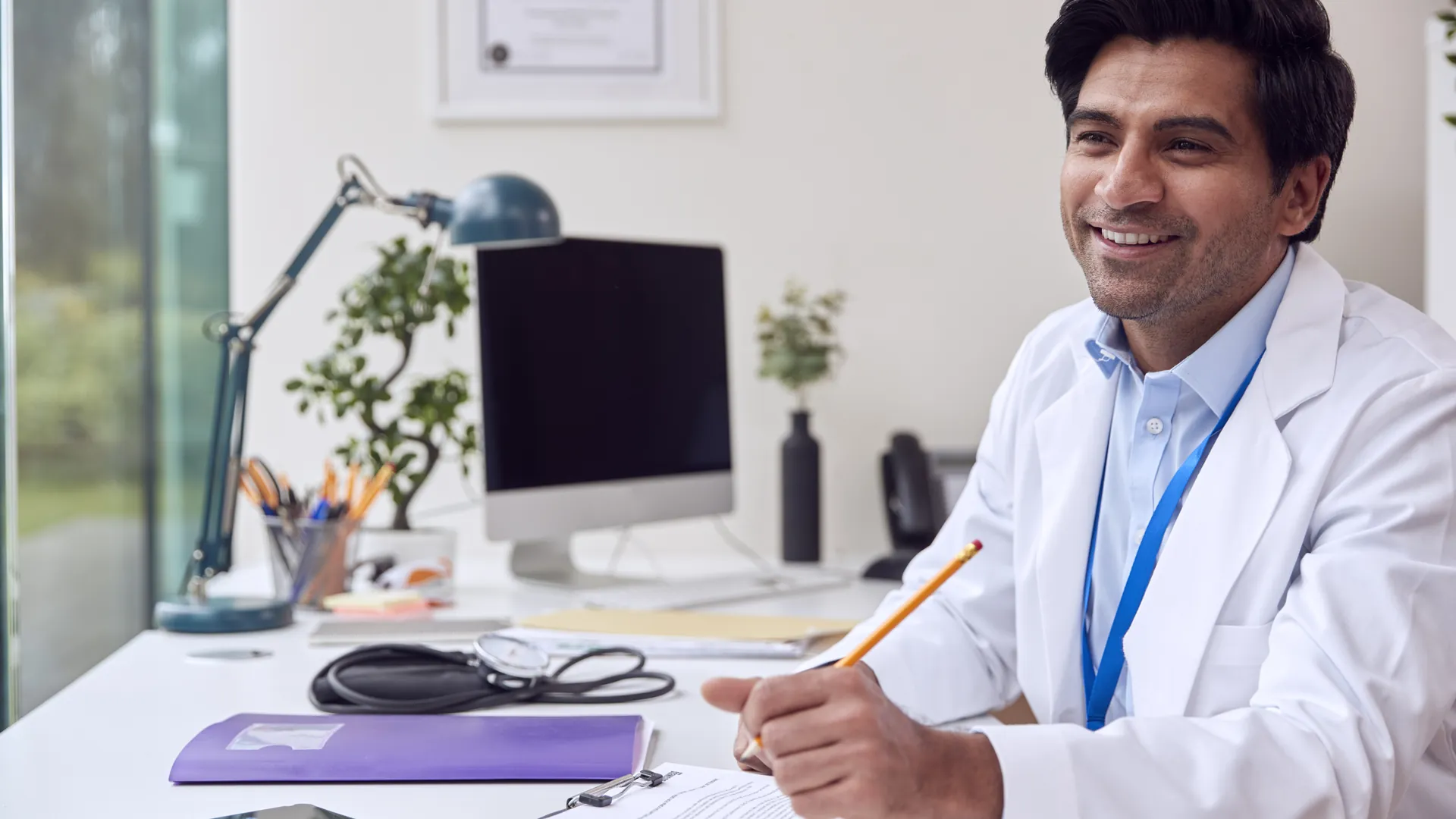 A smiling doctor in a white coat holds a pencil at a tidy desk with a computer monitor, stethoscope and a purple folder.