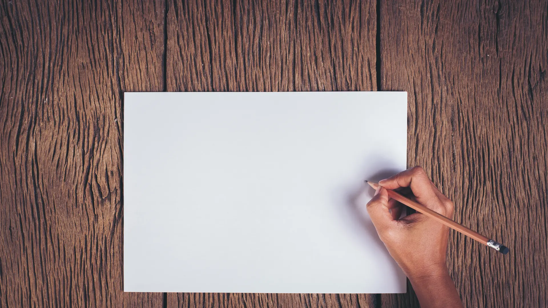 A hand draws with a pencil on a blank sheet of paper lying on a weathered dark wooden table.