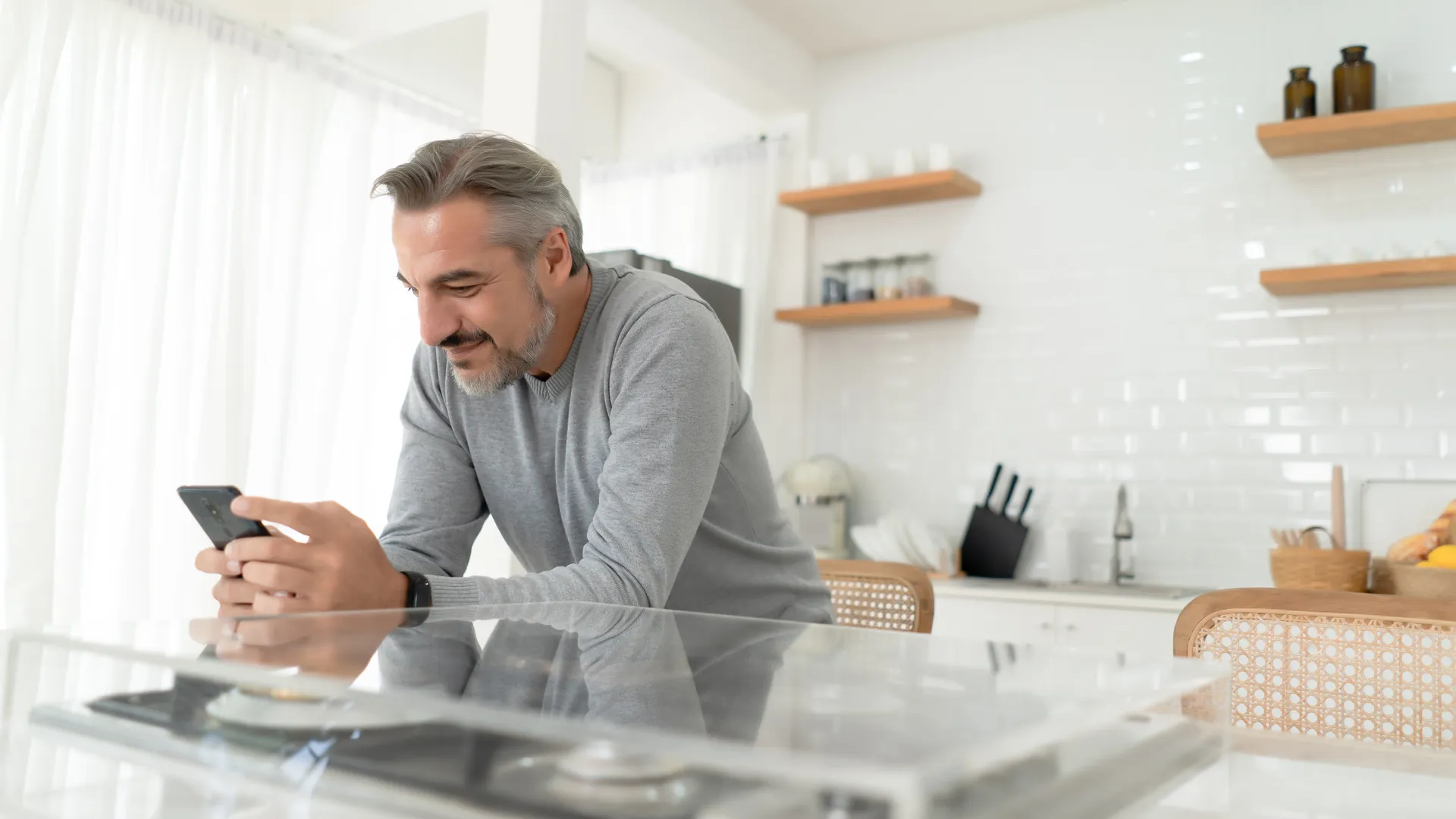 A grey-haired man in a bright kitchen smiles at his phone while leaning on a glass-topped counter.