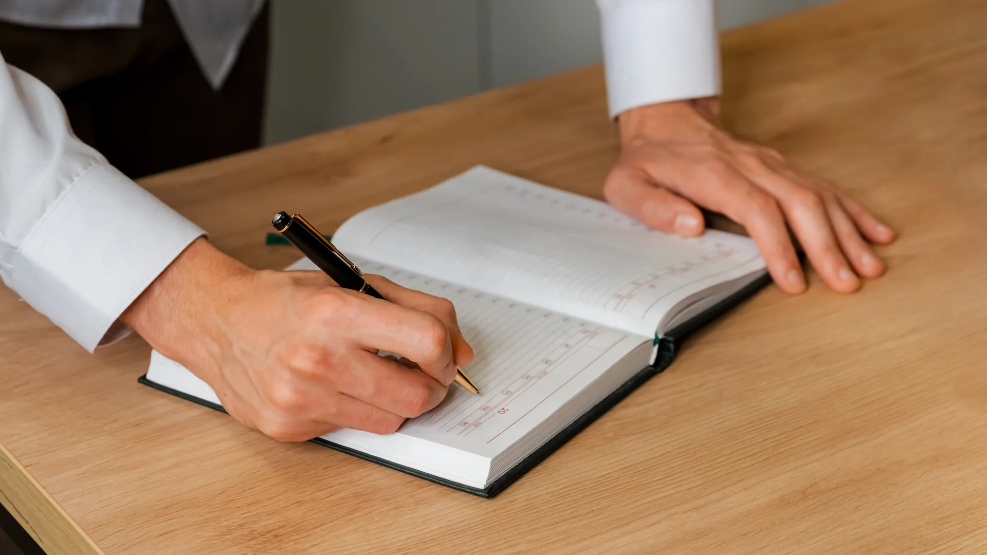 A person writes with a black pen in a diary on a light wooden desk.