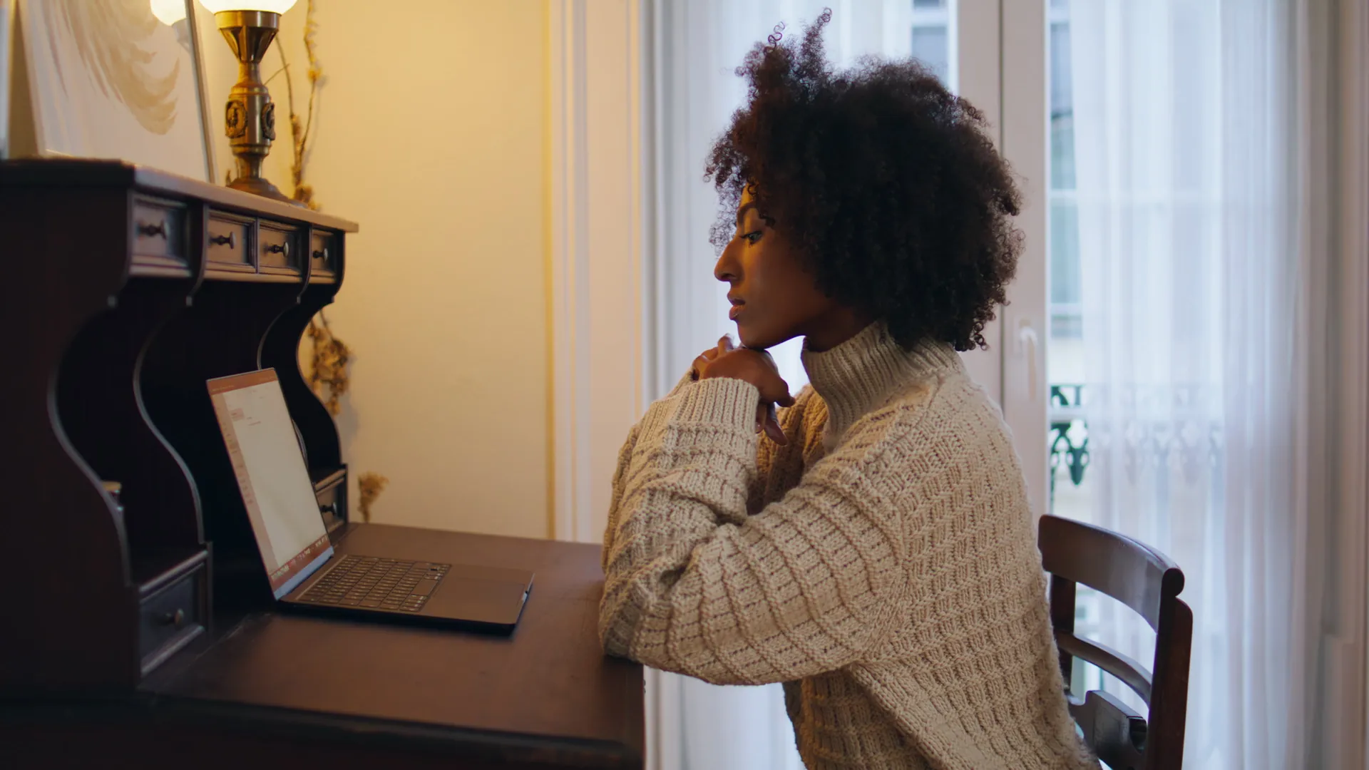 A woman with curly hair rests her chin on her hand as she looks at a laptop on an old wooden desk in a warm room.