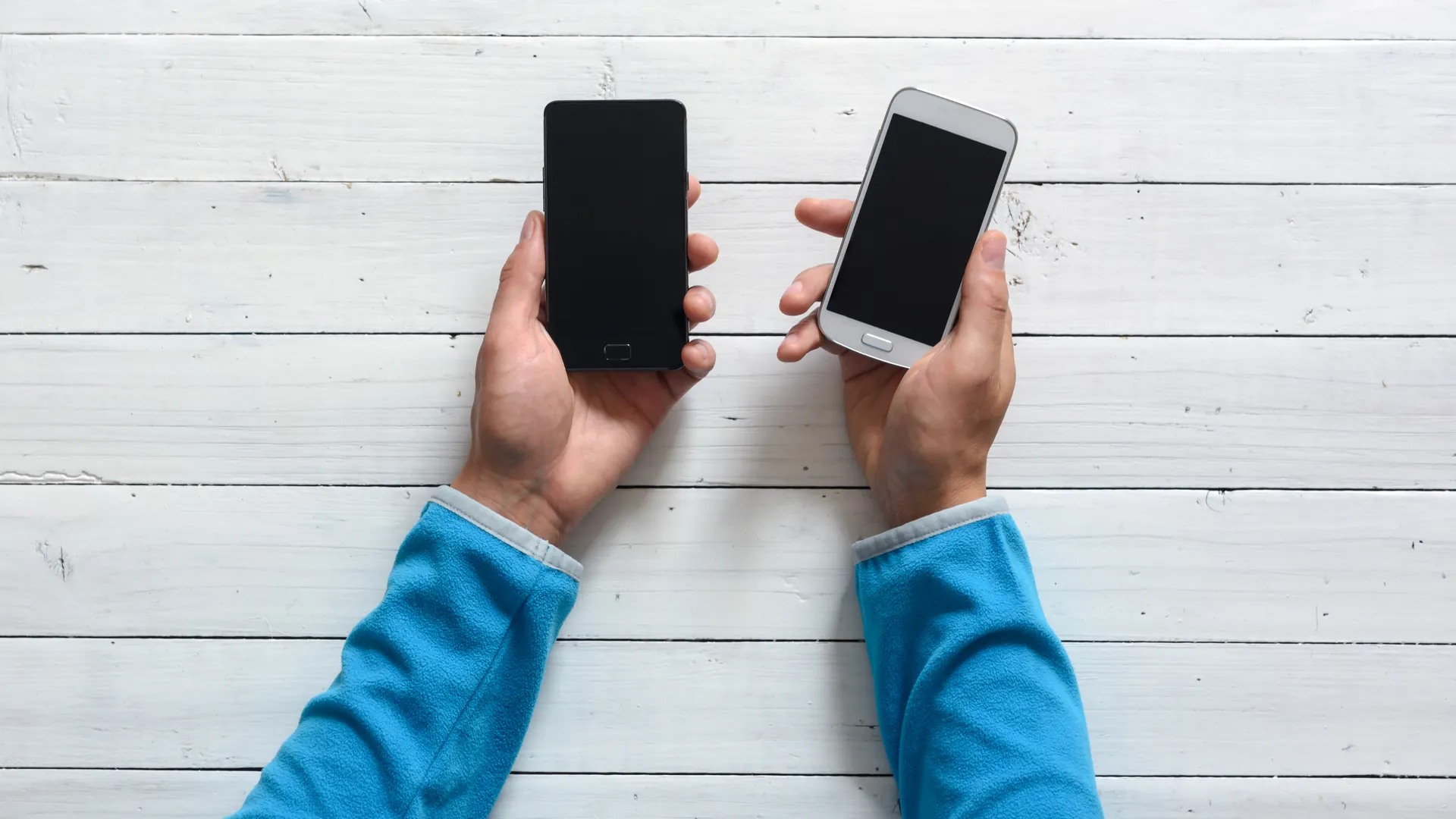 Two hands seen from above, each holding a smartphone, over a white-painted wooden table.