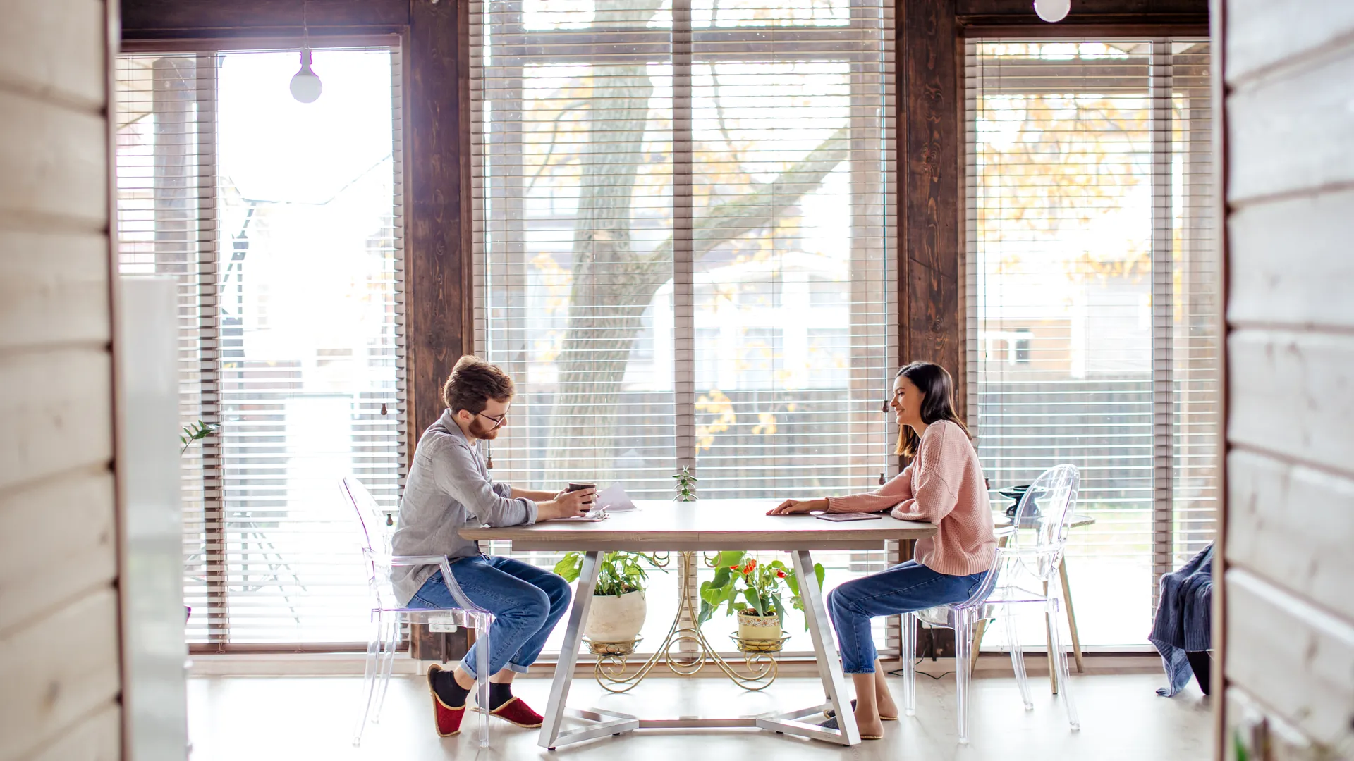 A man and a woman sit opposite each other at a white table by large windows, talking as they look over notes.