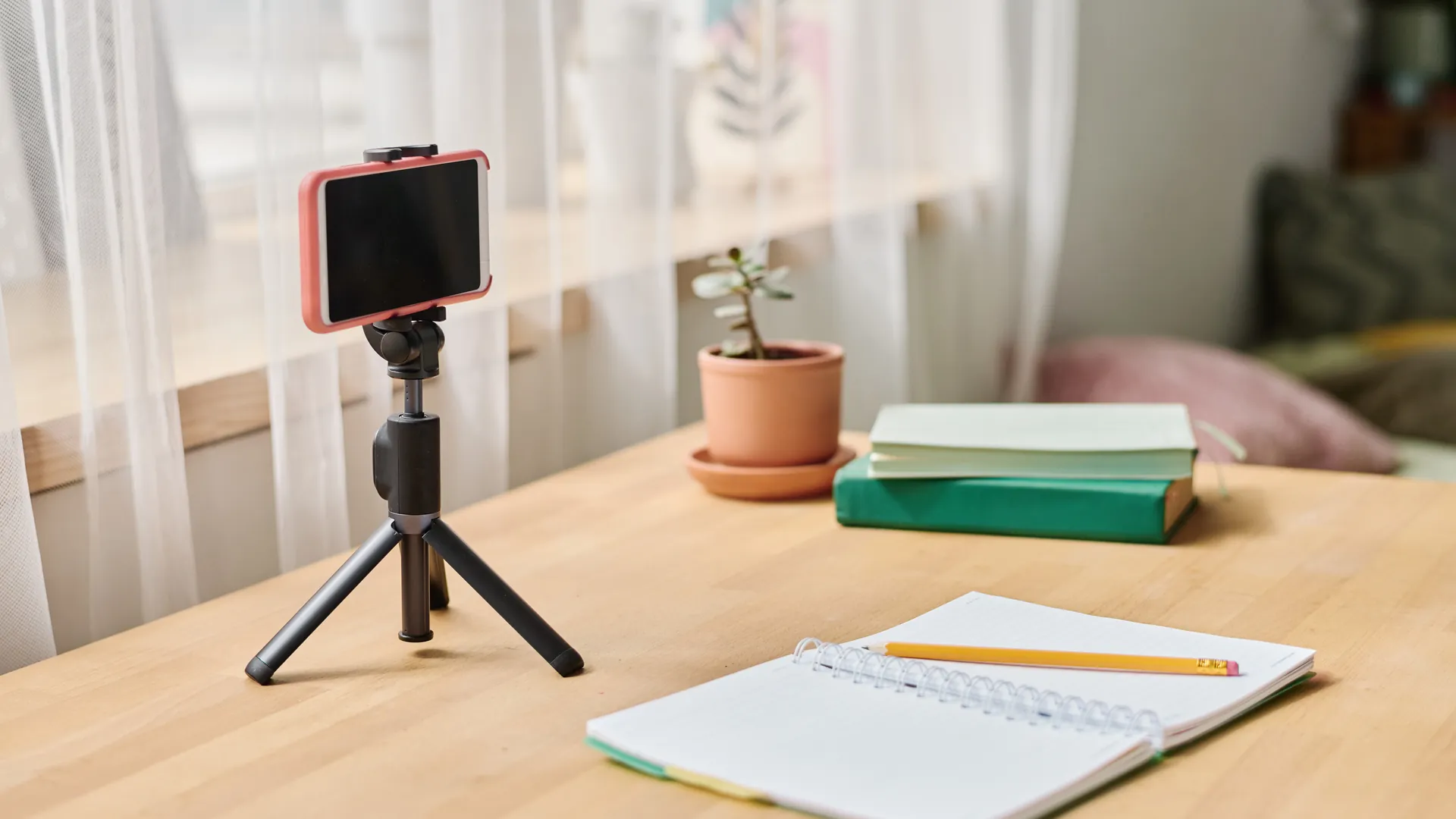 A smartphone on a small tripod beside a notebook, pencil and books on a light wooden desk.