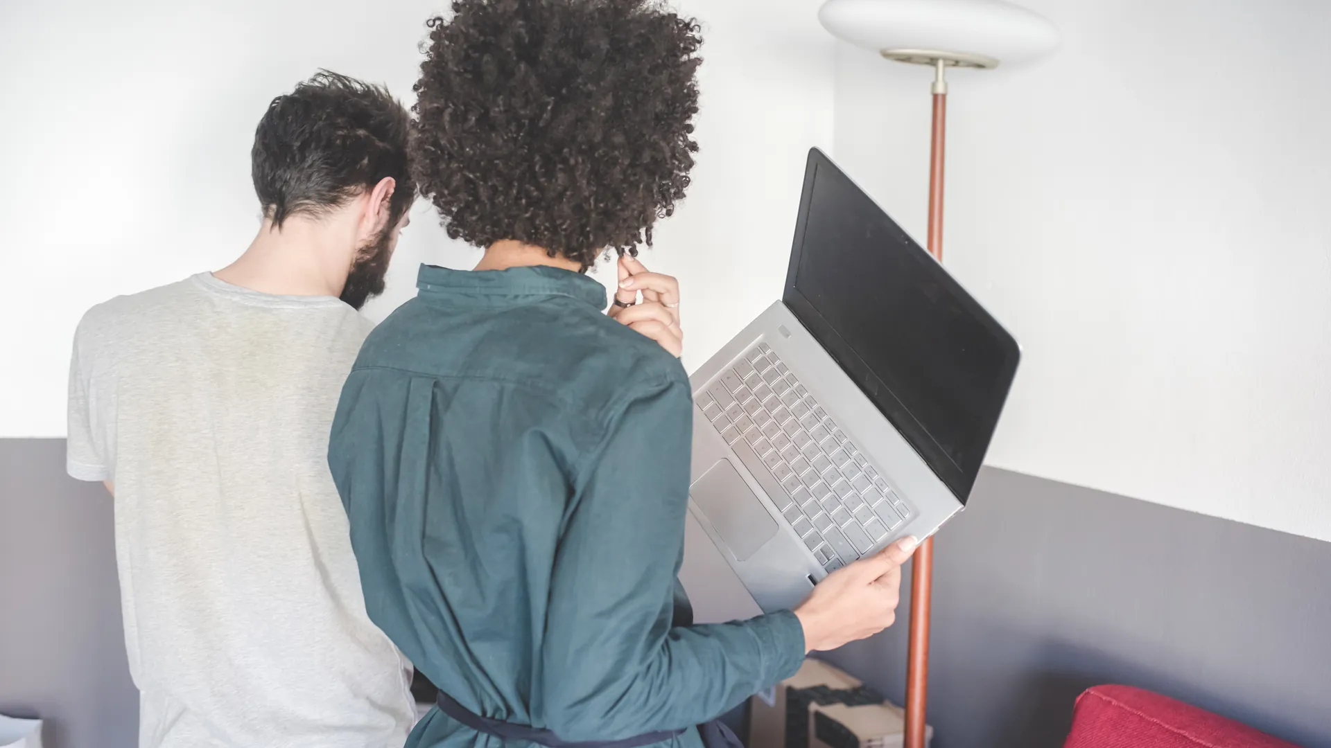 Two people with their backs to the camera stand together looking at a laptop that one of them holds.