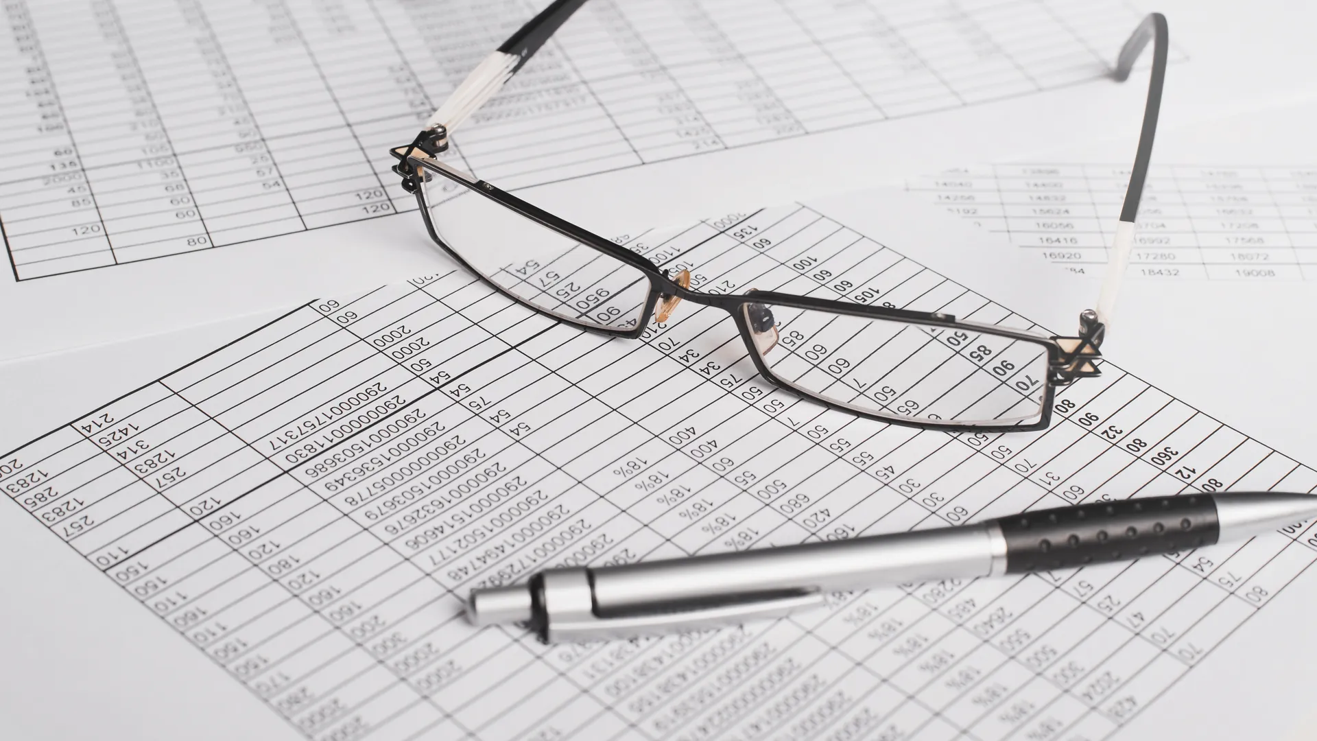 A pair of glasses and a silver pen resting on printed spreadsheets filled with columns of figures.