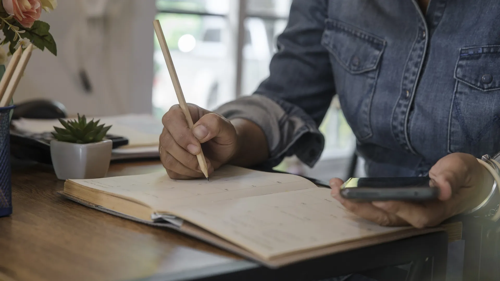 A person in a denim shirt writes in a diary with a pencil while holding a phone at a wooden desk.