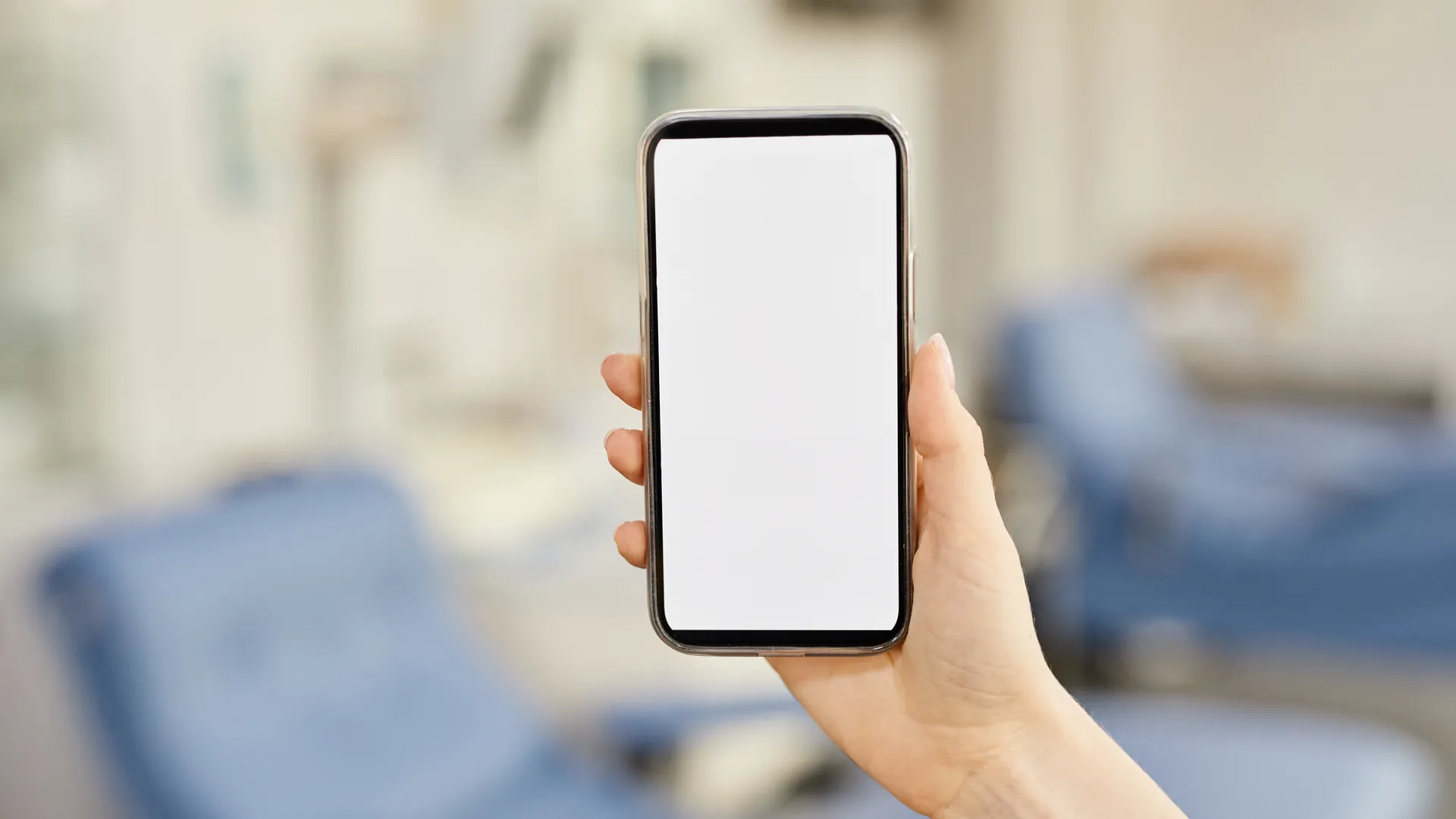 A hand holds a smartphone with a blank screen in front of a softly blurred clinic room with blue chairs.