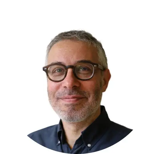 A middle-aged man with short grey hair, light skin, and a beard smiles gently. He wears round dark-rimmed glasses and a dark button-up shirt. Ideal for a healthcare digital marketing agency, the photo is circular on a plain white background.