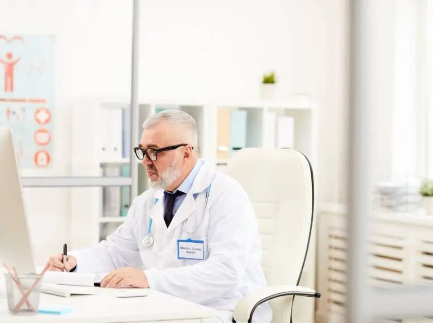 An older male doctor with grey hair and beard, wearing glasses, a white coat, and a stethoscope, sits at a desk in a bright office. He writes on a notepad whilst researching digital marketing healthcare on his computer.