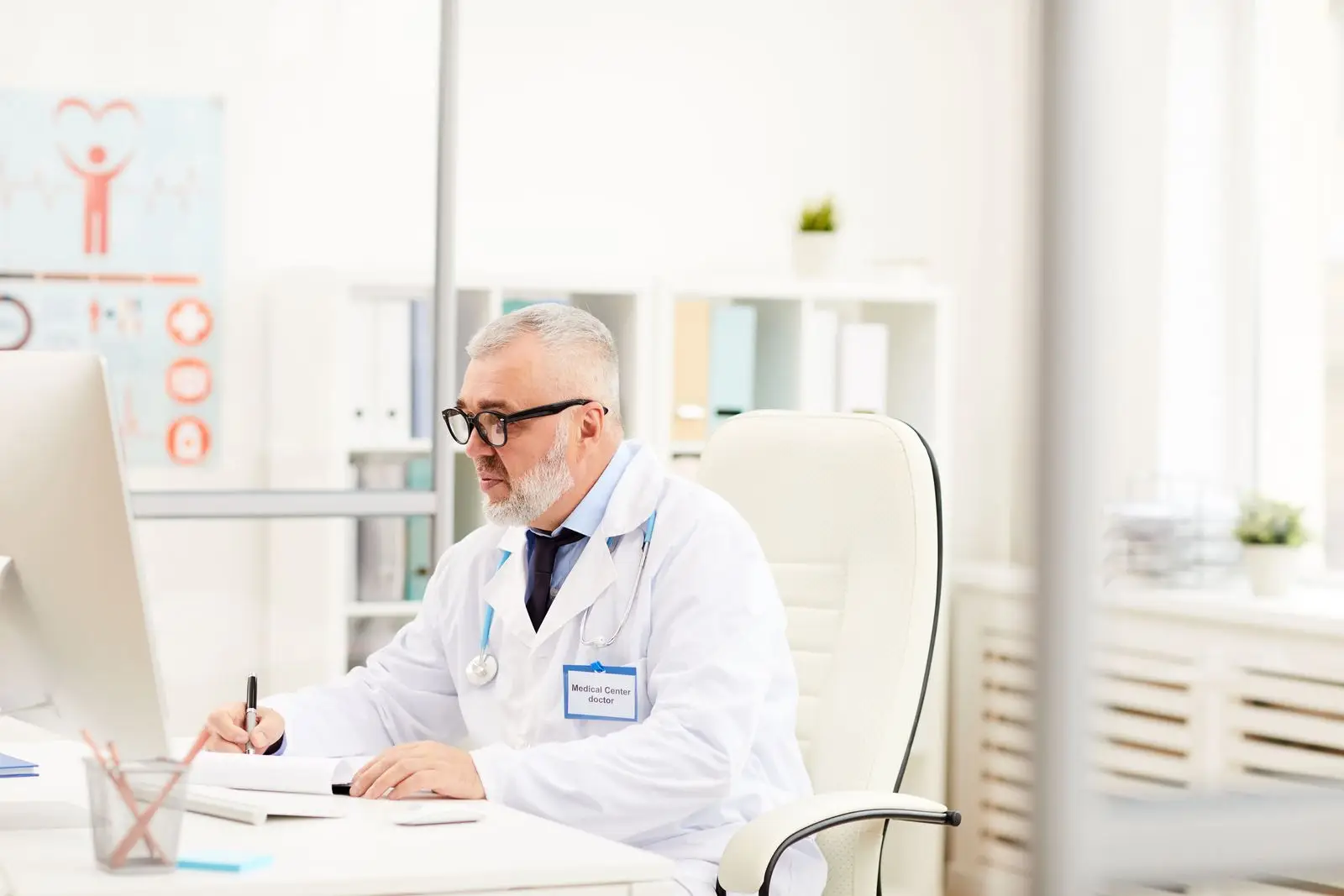 An older male doctor with grey hair and beard, wearing glasses, a white coat, and a stethoscope, sits at a desk in a bright office. He writes on a notepad whilst researching digital marketing healthcare on his computer.