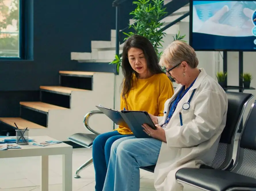 A woman in a yellow jumper sits in a waiting area with a doctor, reviewing documents. Behind them are stairs, plants, a digital medical screen, and brochures—highlighting the modern touch of a healthcare digital marketing agency.