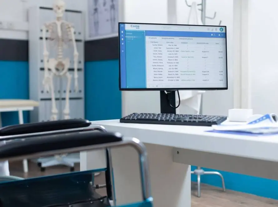 A computer monitor on a white desk displays medical records in a modern doctor’s office. An empty chair faces the desk. In the background, a skeleton model stands near blue and white walls—perfect for a healthcare digital marketing agency shoot.