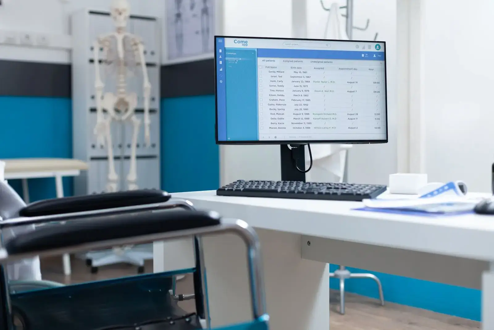 A computer monitor on a white desk displays medical records in a modern doctor’s office. An empty chair faces the desk. In the background, a skeleton model stands near blue and white walls—perfect for a healthcare digital marketing agency shoot.