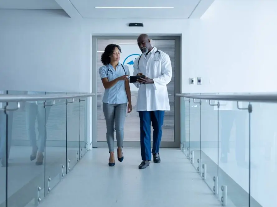 A male doctor in a white coat and a woman in business casual attire, possibly a healthcare SEO consultant, walk down a bright hospital corridor, discussing information on a clipboard. Glass railings line the hallway and a medical symbol is visible on a door behind them.