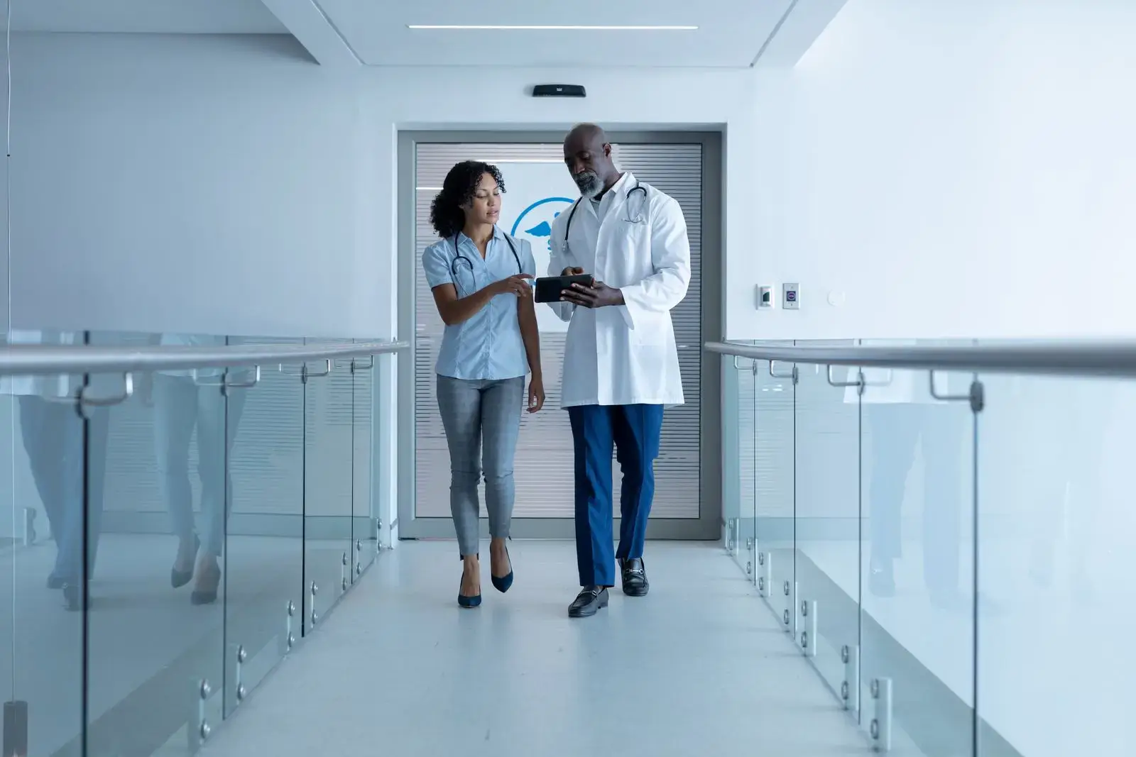 A male doctor in a white coat and a woman in business casual attire, possibly a healthcare SEO consultant, walk down a bright hospital corridor, discussing information on a clipboard. Glass railings line the hallway and a medical symbol is visible on a door behind them.
