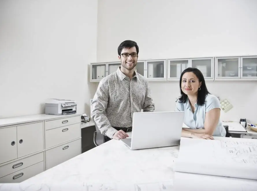 A man and a woman sit at a desk in a bright office with white cupboards and a printer. The man stands, smiling, in front of an open laptop, while the woman sits beside him, also smiling—typical teamwork at a healthcare digital marketing agency.