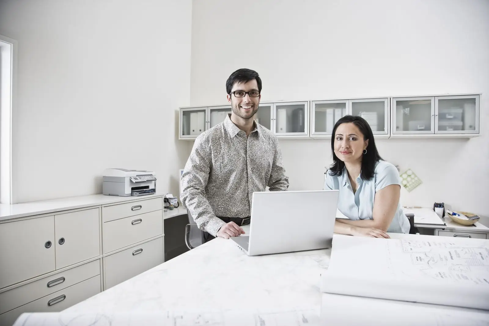 A man and a woman sit at a desk in a bright office with white cupboards and a printer. The man stands, smiling, in front of an open laptop, while the woman sits beside him, also smiling—typical teamwork at a healthcare digital marketing agency.