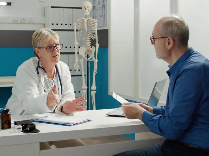 A female doctor in a white coat speaks to an older male patient across a desk in a medical office. Medical documents, a stethoscope, and a pill bottle are on the desk—ideal imagery for a digital marketing healthcare campaign. Shelves and a skeleton model are in the background.