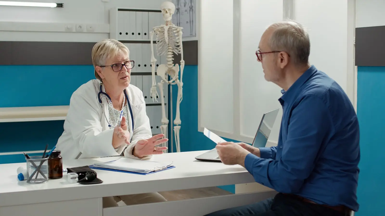 A female doctor in a white coat speaks to an older male patient across a desk in a medical office. Medical documents, a stethoscope, and a pill bottle are on the desk—ideal imagery for a digital marketing healthcare campaign. Shelves and a skeleton model are in the background.