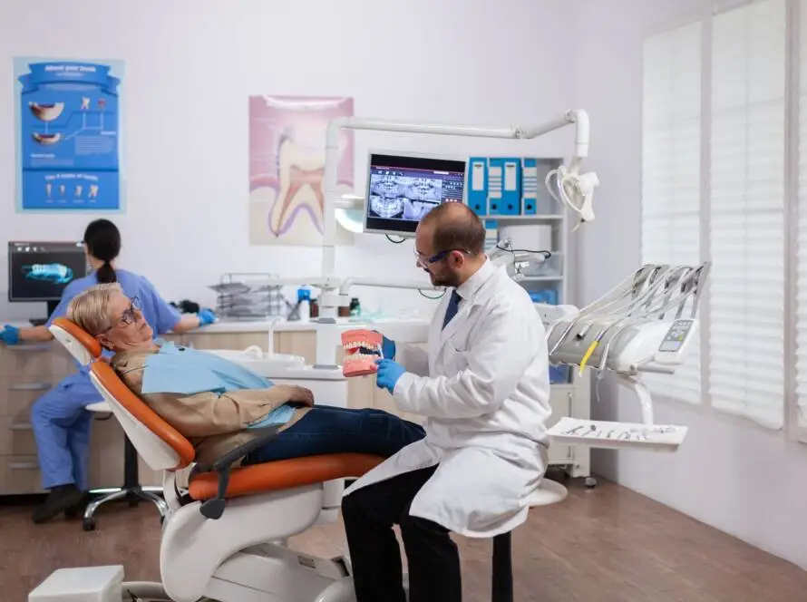 A dentist in a white coat shows a dental model to an elderly patient in an orange chair. Another professional works nearby. The bright room, featured by a medical marketing agency, has posters, dental instruments, and a large window.