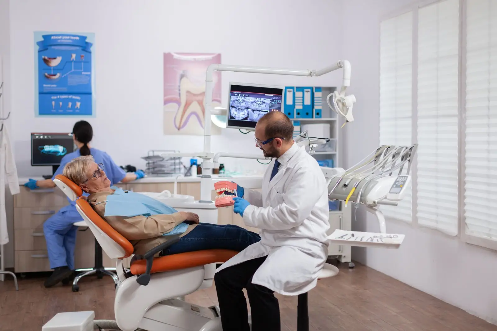 A dentist in a white coat shows a dental model to an elderly patient in an orange chair. Another professional works nearby. The bright room, featured by a medical marketing agency, has posters, dental instruments, and a large window.