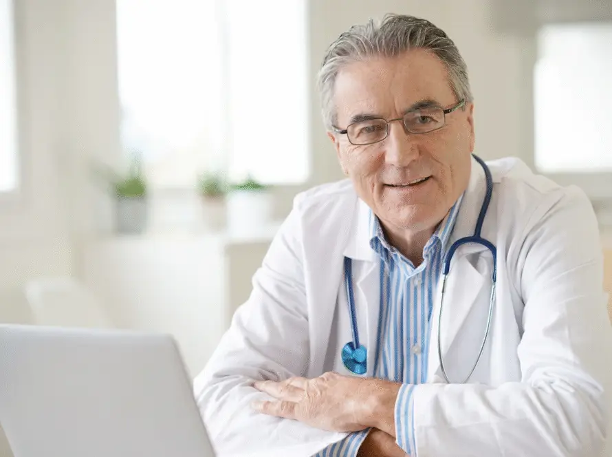 Smiling older male doctor at desk with stethoscope and laptop, featured on www.pulsedigital.health in a bright, modern office.