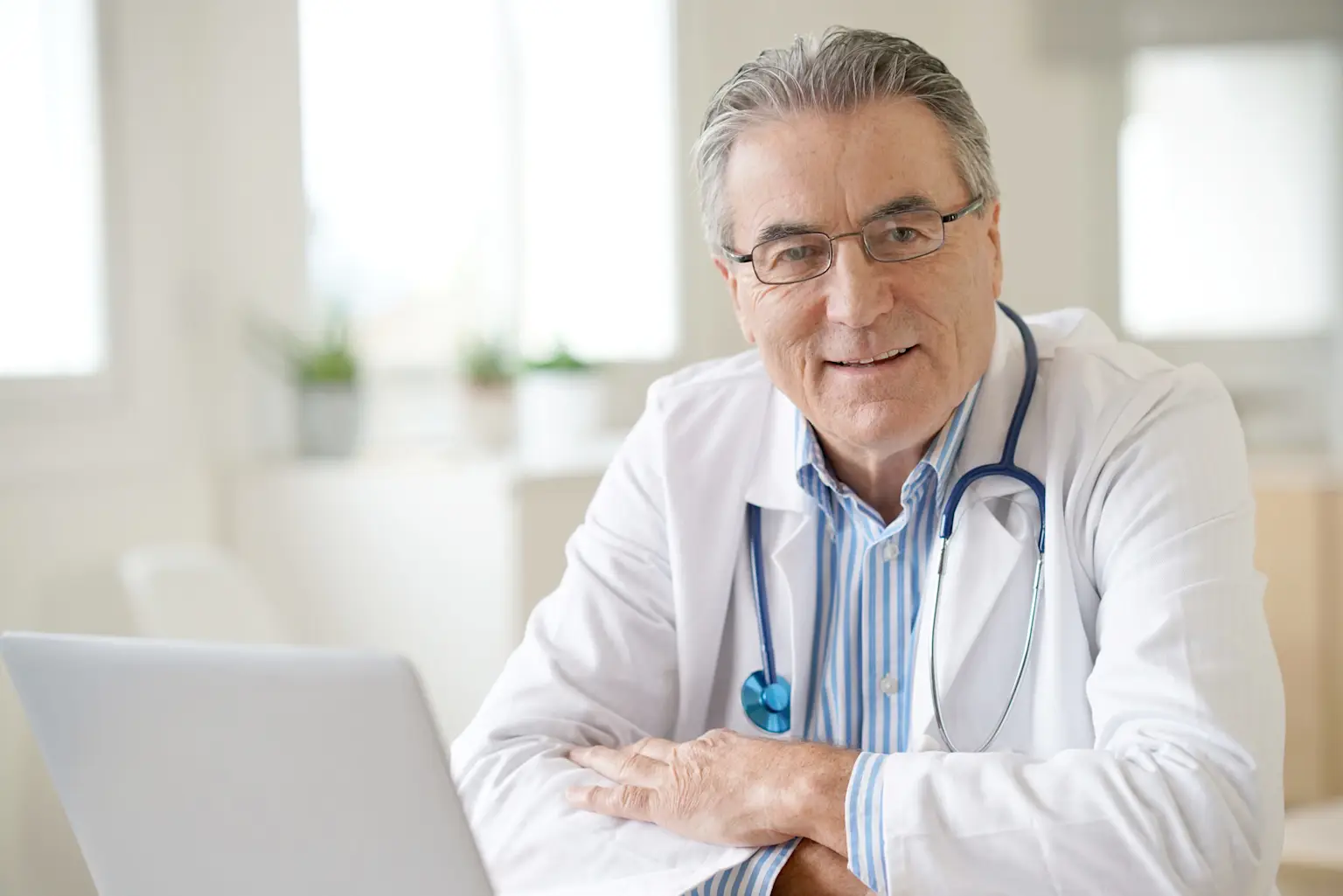 Smiling older male doctor at desk with stethoscope and laptop, featured on www.pulsedigital.health in a bright, modern office.
