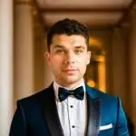 A young man with short brown hair and blue eyes stands in a corridor, dressed smartly for an event hosted by a leading healthcare digital marketing agency. He looks directly at the camera with a neutral expression.
