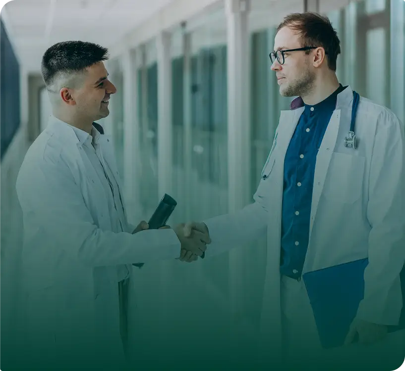 Two male doctors in white lab coats shake hands in a bright corridor. One holds a rolled-up document and smiles, while the other, with glasses and a stethoscope, holds a blue clipboard—perhaps discussing insights from a healthcare digital marketing agency.
