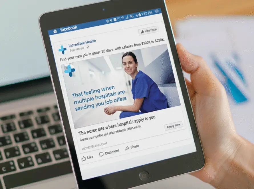 A person holds a tablet displaying a Facebook advert for a nurse job site, created by a leading medical marketing agency. The advert features a smiling nurse in blue scrubs with the text, “That feeling when multiple hospitals are sending you job offers.”.